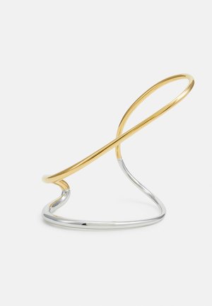 Support en métal avec une finition dorée et argentée, présentant un design courbé et minimaliste. Texture lisse, forme de boucle ouverte et éléments entrelacés.