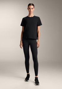 Žena stojící vzpřímeně, oblečená v černém tričku s krátkým rukávem, černých legínách a černých teniskách na jednobarevném béžovém pozadí.