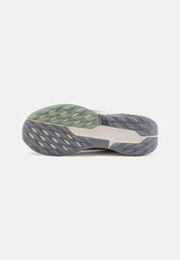 Suola di scarpe da trail running in diverse tonalità di verde, grigio e crema. Presenta tasselli multidirezionali per una migliore aderenza e un materiale in gomma resistente.
