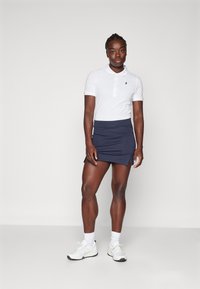 Weißes Polo-Shirt mit kurzen Ärmeln und navyfarbenem Rock mit seitlichem Schlitz. Weiße Sportschuhe und knöchellange Socken vervollständigen das Outfit.