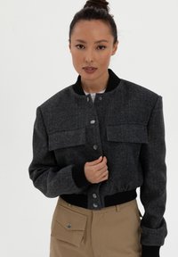 Harmaa herringbone-takki, jossa on kaksi etutaskua, nepparikiinnitys ja mustat ribattuja hihansuut ja kaulus, yhdistettynä khaki-housuihin.