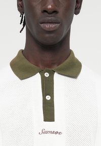 Άσπρο πλεκτό πόλο με γιακά σε ελιά πράσινο, δύο κουμπιά και κεντημένο "Samsoe" σε μπορντό. Υφή υφάσματος με οπές.