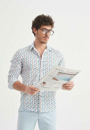 Chemise blanche à manches longues avec des motifs ovales verticaux multicolores ; le modèle tient un journal, porte des lunettes et a les cheveux courts et bouclés.