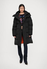 Giacca puffer nera con cappuccio, dotata di chiusura con zip e bottoni a pressione. Fodera interna arancione. Indossata sopra jeans scuri e una maglietta colorata.