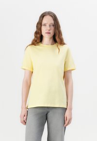 T-shirt en coton jaune avec un col rond et des manches courtes. Présente une coupe décontractée et une texture lisse. Associé à un jean en denim gris.