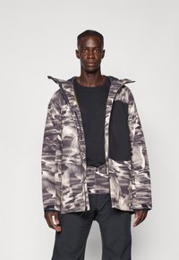 Chaqueta con patrón de camuflaje que cuenta con capucha, cierre de cremallera y bolsillos. Fabricada con un tejido duradero, principalmente en tonos de gris y blanco.
