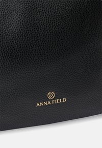 Black textured leather bag with gold logo "ANNA FIELD" at the bottom, featuring a minimalist design and no visible hardware.