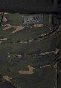Gröna kamouflagedbyxor med en ficka i lappstil, bruna accenter och en svart läderetikett i midjan präglad med "ONLY." Mjuk, slitstark tyg med sömnadsdetaljer.
