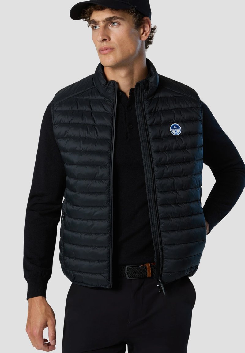 Chaleco de plumas negro con cremallera frontal, textura acolchada horizontal y un parche de logo redondo en el lado izquierdo del pecho. Lleva puesto un top negro de manga larga.