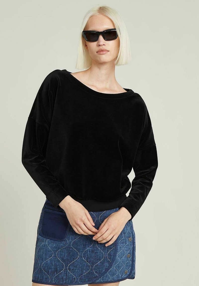 Schwarzes Samtoberteil mit langen Ärmeln, lockerer Passform und weitem Ausschnitt, kombiniert mit einem blauen Jeansrock, der über eine blaue Tasche und wellenförmige Nähte verfügt.