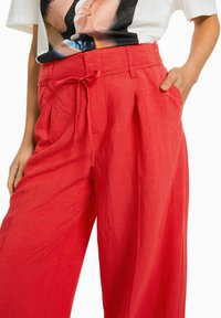 Rote Leinenhose mit lockerer Silhouette, gefälteltem Frontbereich, verstellbarem Kordelzugbund und seitlichen Taschen, ausgestattet mit einer glatten, leichten Textur.