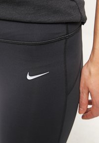 Leggings deportivos negros de ajuste ceñido, con un logo blanco de Nike, textura suave y costuras visibles a lo largo de las costuras.