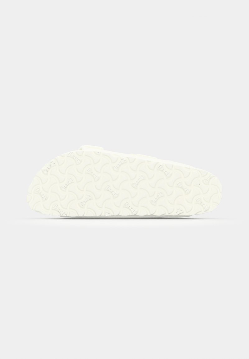 Zapato blanco sin cordones con una suela de goma texturizada que presenta un patrón ondulado, superficie superior lisa y diseño minimalista.