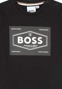 T-shirt en coton noir avec un grand graphisme blanc portant le mot "BOSS" et des motifs de texte. Encolure ronde avec des boutons pression.