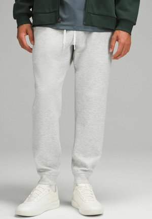 Pantalons de survêtement gris en coton avec une taille élastique et des cordons de serrage blancs, associés à des baskets blanches, sur un fond neutre.