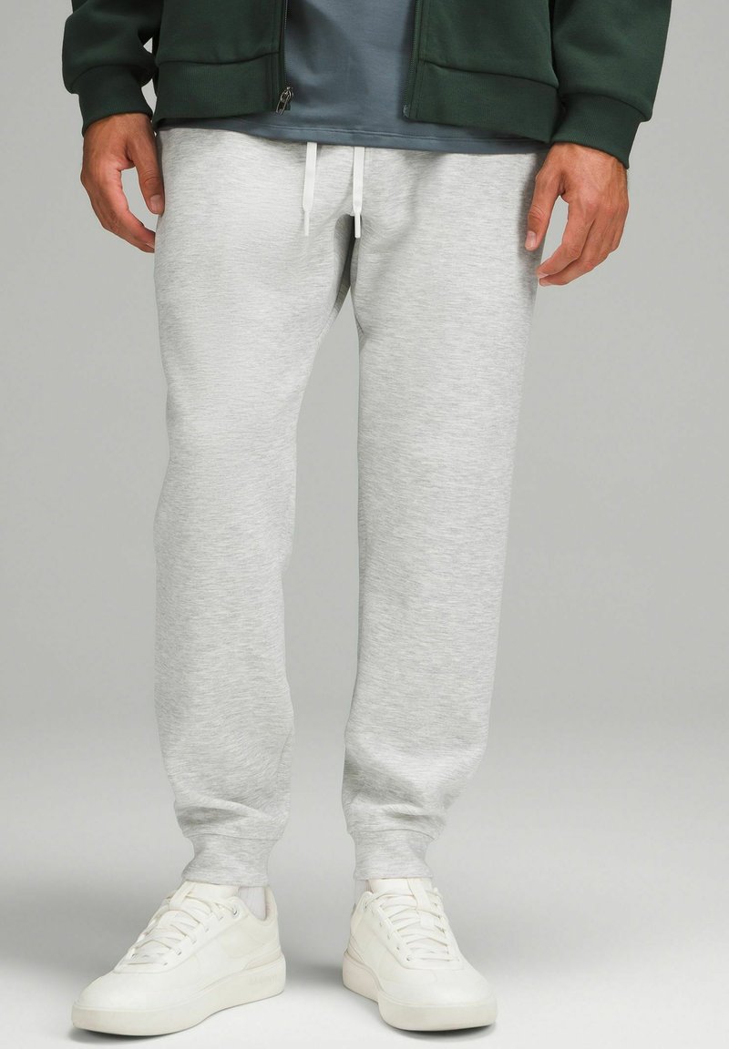 Grå bomullssweatpants med elastisk midjeband och vita snören, i kombination med vita sneakers, mot en neutral bakgrund.