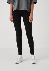 Leggings noirs en tissu extensible, design ajusté, texture lisse, associés à des baskets de sport blanches et un haut oversized gris.