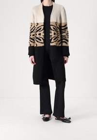 Długi kardigan w kolorach kremowym, beżowym i czarnym. Posiada wzór zwierzęcy, otwarty przód oraz ściągacze przy mankietach. Noszony na czarny strój i buty.