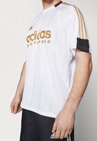 Vit Adidas-tröja med guldlogotyp, vertikala ränder och svarta detaljer på ärmarna. Har rund halsringning och avslappnad passform.