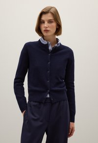 Cardigan blu navy in morbido tessuto a maglia con collo rotondo, chiusura a bottoni e polsini a costine, indossato sopra una camicia a righe blu e bianche.