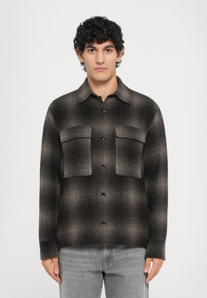 Man with curly dark hair wearing a brown and black plaid button-up shirt with chest pockets and light gray jeans, facing forward.