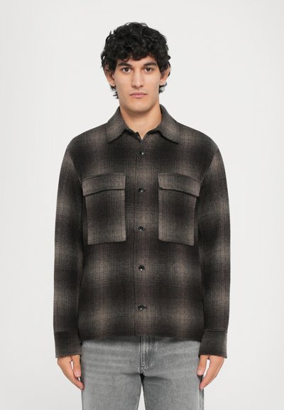 Homme aux cheveux bouclés et foncés portant une chemise à carreaux marron et noir à boutons avec des poches sur la poitrine et un jean gris clair, de face.