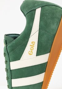 Grüner Wildleder-Sneaker mit weißen Streifen und goldenem Logo. Verfügt über eine strukturierte Oberfläche und eine Gummisohle mit ausgeprägten Rillen für bessere Traktion.