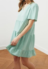 Světle zelené šaty s krátkým rukávem, v hlubokém výstřihu, s vrstveným designem a volánkovým lemem. Vyrobené z měkké, texturované látky.