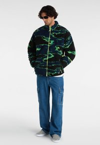 Fluffy zwarte jas met groene golfpatronen en een voorrits. Gepaard met blauwe cargobroeken en geruite instappers.