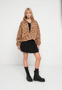 Καφέ φλις μπουφάν με λεοπάρ μοτίβο, ολόκληρο φερμουάρ και ψηλό γιακά, σε συνδυασμό με μαύρη φούστα και μαύρες χοντρές μπότες.