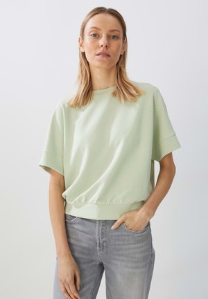 Mintgrüner Pullover mit kurzen Ärmeln, lockerer Passform und gerafftem Saum, kombiniert mit hellgrauen Jeans. Glatte Textur, minimalistisches Design.