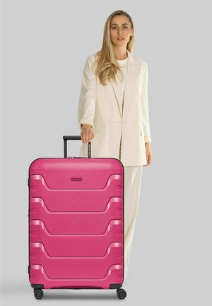 Femme blonde en costume beige debout, tenant la poignée d'une grande valise rigide rose à quatre roues sur fond gris.