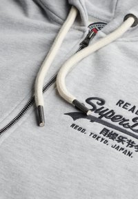 Sudadera gris con cremallera, hecha de tela suave, que cuenta con cordones color crema con acentos metálicos y texto estilizado en negro en el pecho.