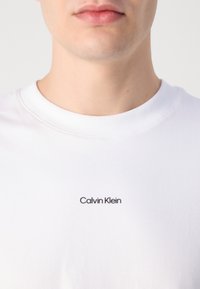 Biela bavlnená tričko s kratkým rukávom a okrúhlym výstrihom, na prednej strane s malým čiernym logom "Calvin Klein", hladká štruktúra a klasický strih.