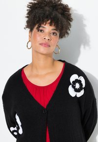 Schwarzer Cardigan mit V-Ausschnitt aus strukturiertem Strickstoff. Mit weißer floraler Stickerei und einem einzelnen Knopfverschluss. Getragen über einem roten Oberteil.