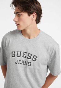 Pilkas medvilnės t-shirt'as su juodu spausdintu logotipu, ant kurio užrašyta "GUESS JEANS". Trumpomis rankovėmis ir apvaliu iškirpimu, laisvas pjūvis.