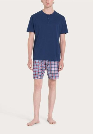 Giovane uomo che indossa una maglietta henley blu navy a maniche corte e pantaloncini a quadri rossi, bianchi e blu, in piedi a piedi nudi su uno sfondo semplice.