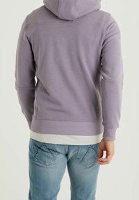 Haut à capuche violet en tissu doux avec un cordon de serrage, des poignets et un ourlet côtelés, superposé sur une chemise blanche, associé à un jean bleu clair.