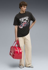 Tricou grafic negru cu un design de mașină de curse, purtat cu pantaloni largi de culoare crem. Ținând o geantă roșie strălucitoare cu accente albe.