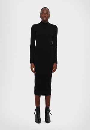 Robe en tricot noire à manches longues avec un col haut, silhouette ajustée et longueur midi, associée à des bottines. Texture lisse, design minimal.