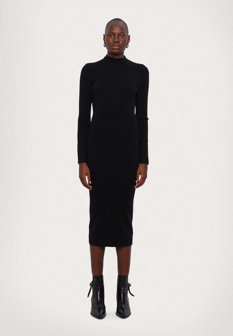 Robe en tricot noire à manches longues avec un col haut, silhouette ajustée et longueur midi, associée à des bottines. Texture lisse, design minimal.