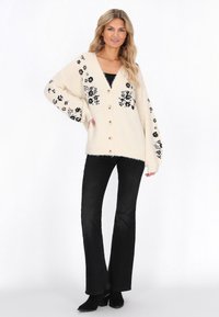 Romantische gebreide cardigan in crème met zwarte bloemenversieringen, knoopsluiting en oversized mouwen. Gecombineerd met zwarte flare jeans en enkellaarsjes.