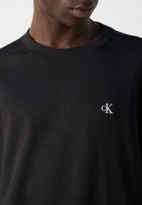 Mann med en svart rundhalset t-skjorte med en liten hvit "cK"-logo brodert på venstre bryst.
