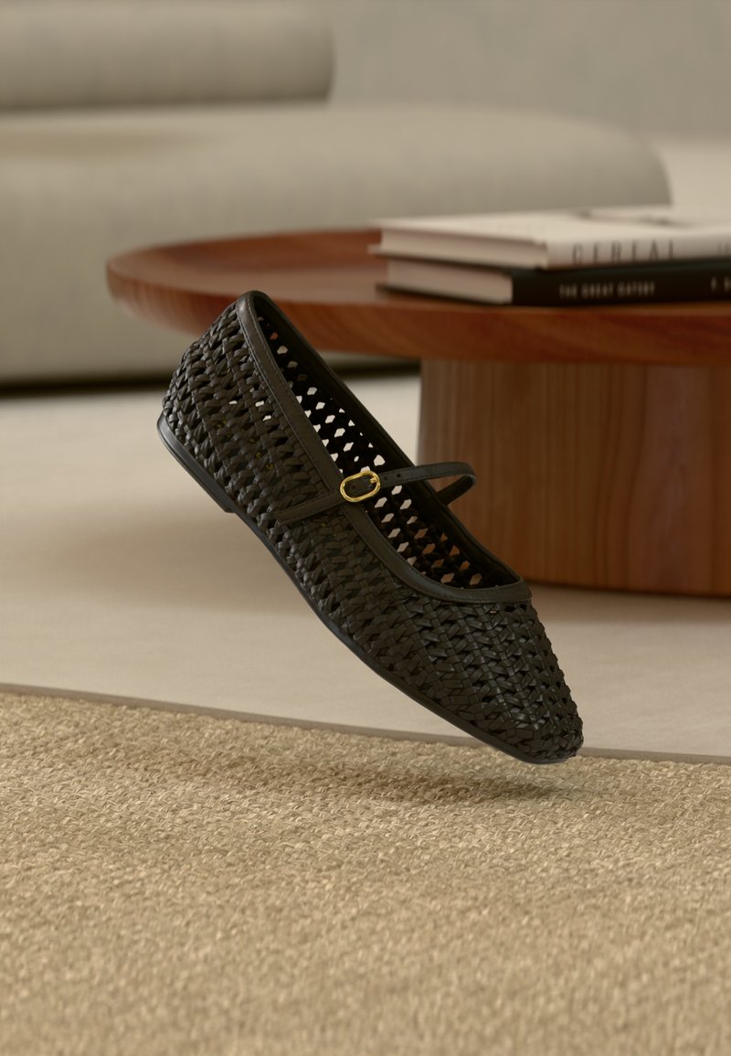 Fekete szövött Mary Jane lapos cipő arany csattal, bézs szőnyeg felett lebegve, háttérben fa dohányzóasztal és világosszürke kanapé.