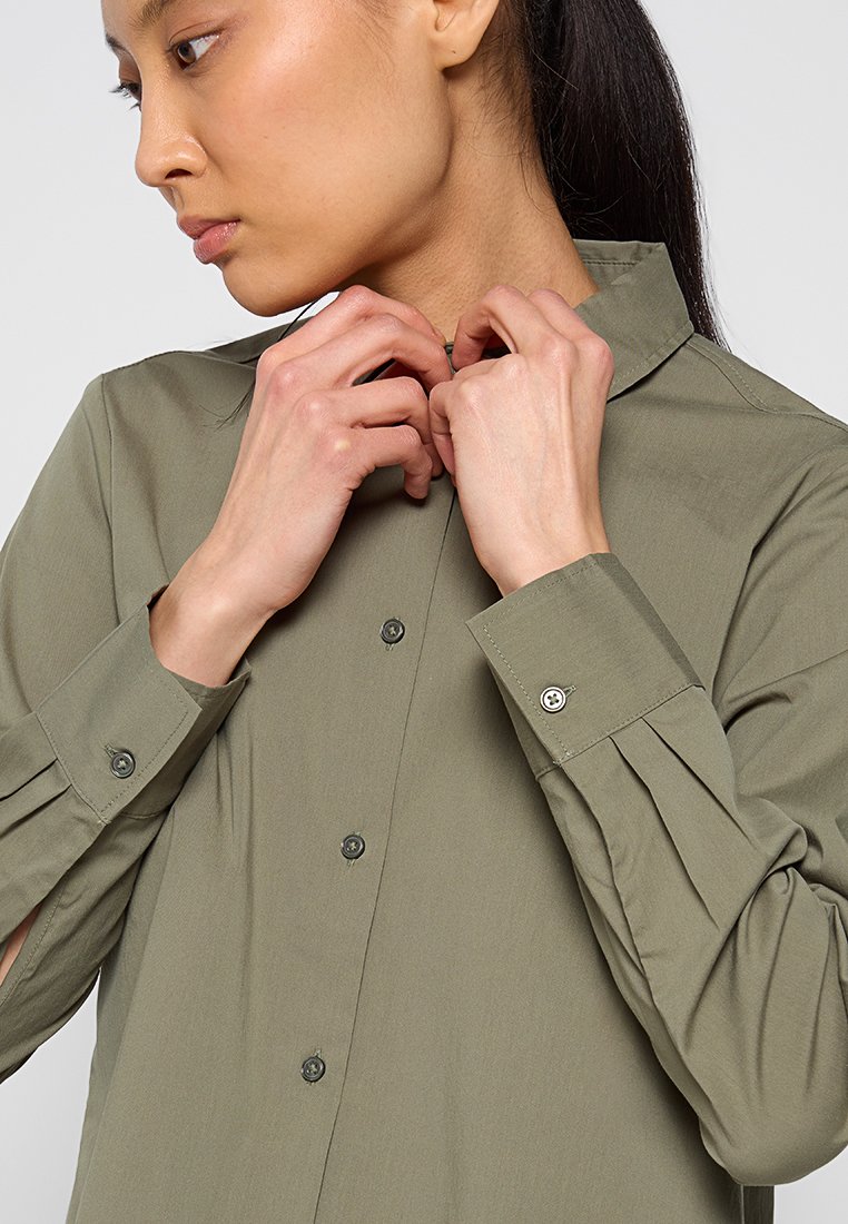 Olivenfarbenes Hemd mit Knopfleiste, spitzem Kragen, langen Ärmeln und gefälteltem Detail an den Schultern. Der Stoff wirkt glatt und leicht.