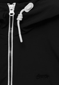 Svart zip-jacka med en texturerad yta. Den har vit dragsko och dragkedjedetaljer, samt en subtil logotyp på framsidan.