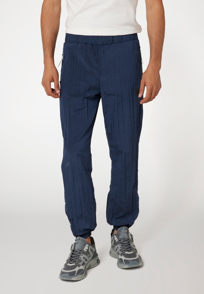 Jūrų laivyno spalvos, lengvi kelnių su tekstūruotu paviršiumi, elastiniu juosmeniu, šoninėmis užtrauktukais ir siaurėjančiais rankogaliais, skirti komfortui ir judrumui.