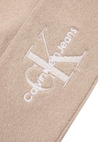 Beżowa dzianina z haftowanym białym logo „CK” oraz napisem „CalvinKleinJeans”. Gładka faktura, subtelny połysk i precyzyjne detale szwów.