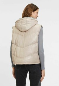 Beige Steppweste mit Kapuze, ausgestattet mit einem gesteppten Zickzackmuster, aus synthetischem Material und einem grauen, ribbed Langarmshirt darunter.