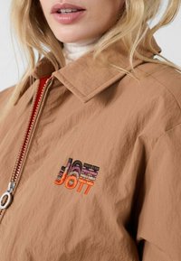 Giacca marrone chiaro con chiusura a zip e colletto a punta. Presenta un logo ricamato multicolore e un tessuto liscio e strutturato.